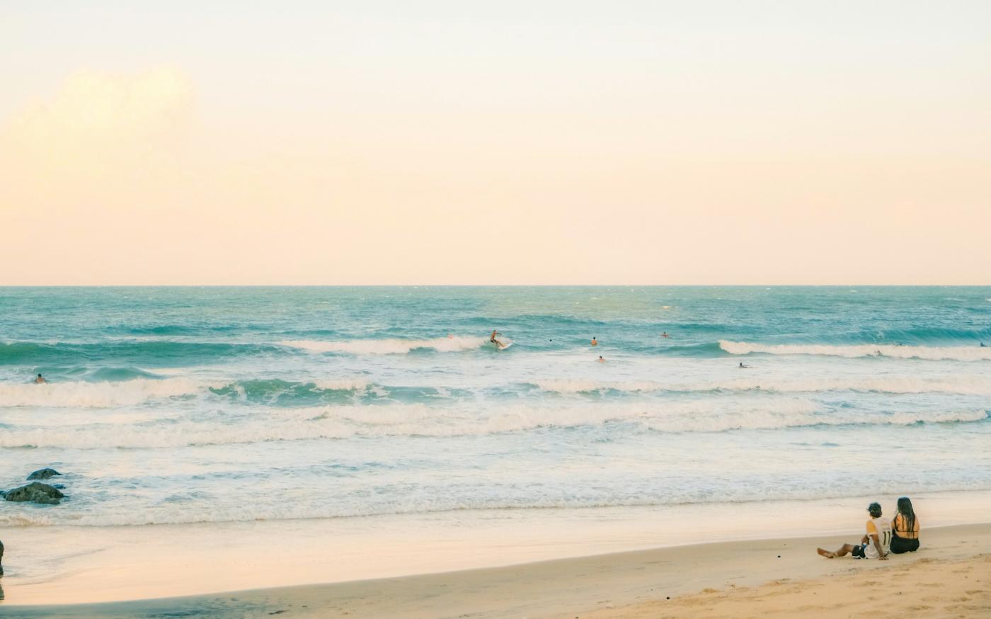 Two people sitting on a sandy beach watching surfers in the ocean by Ivett M courtesy of Unsplash.