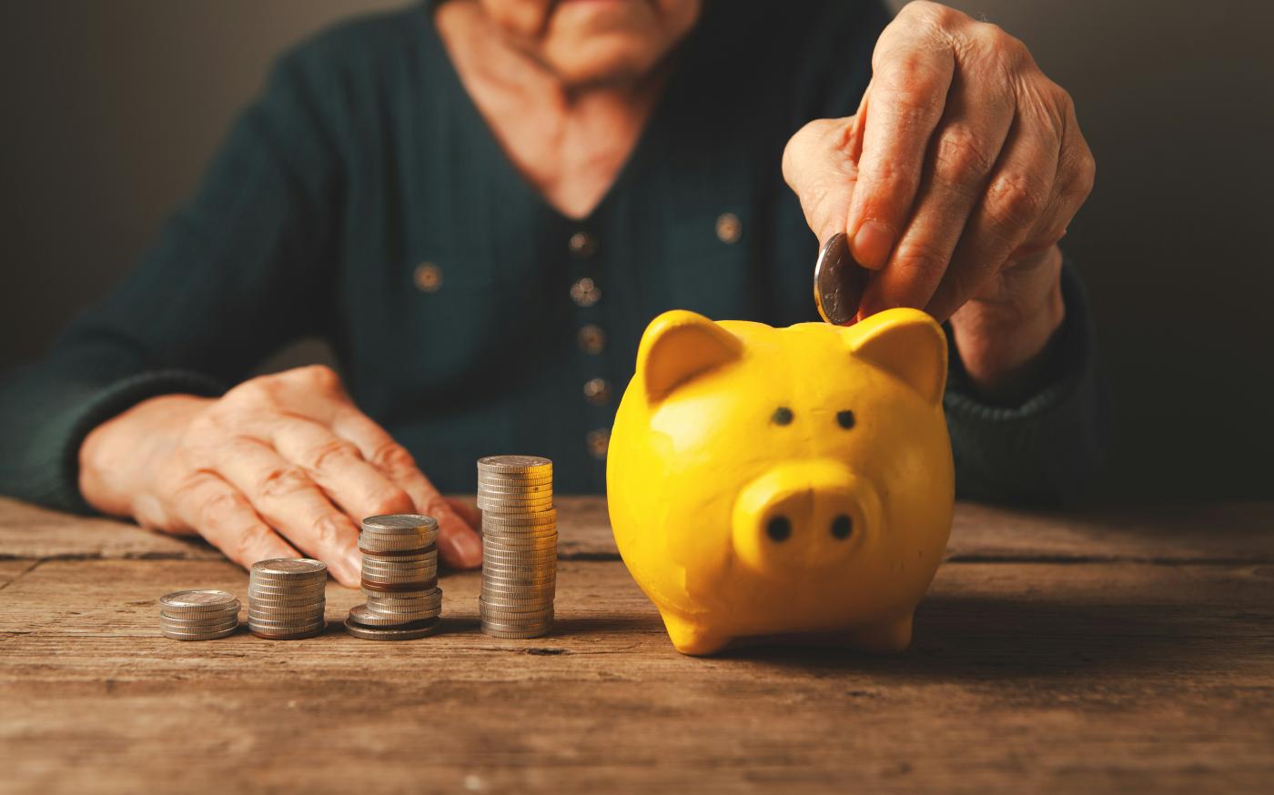 Elderly hands depositing coins into a yellow piggy bank. by Sasun Bughdaryan courtesy of Unsplash.