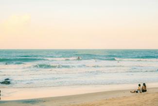 Two people sitting on a sandy beach watching surfers in the ocean by Ivett M courtesy of Unsplash.
